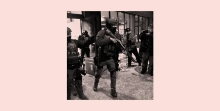 Black-and-white photo of heavily armed law enforcement officers moving through a city sidewalk at night as photographers document the scene.