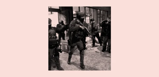 Black-and-white photo of heavily armed law enforcement officers moving through a city sidewalk at night as photographers document the scene.