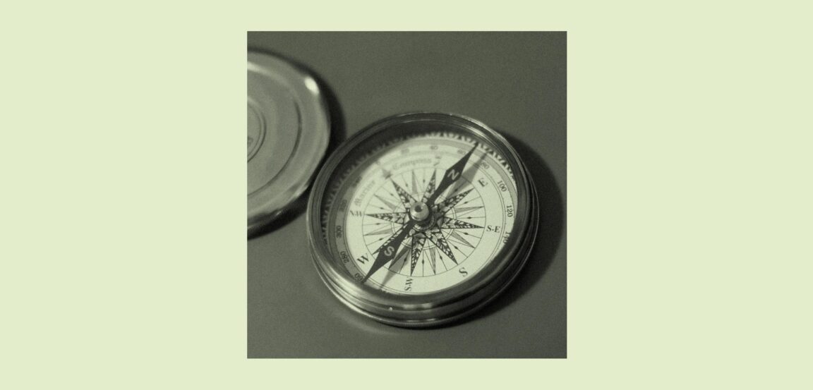 A close-up, black-and-white photograph of an open pocket compass with a decorative wind rose dial, metal casing, and its lid resting beside it on a flat surface. The compass needle points roughly north-northeast.