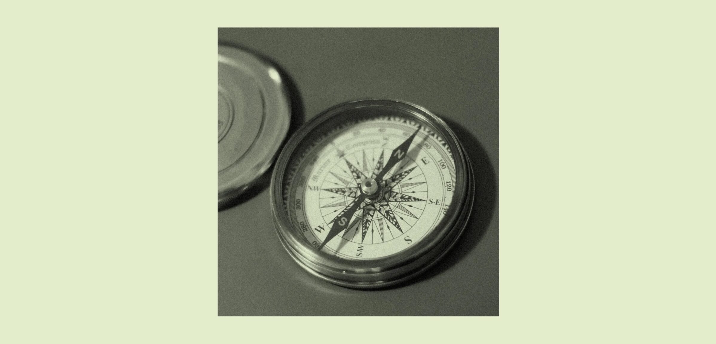 A close-up, black-and-white photograph of an open pocket compass with a decorative wind rose dial, metal casing, and its lid resting beside it on a flat surface. The compass needle points roughly north-northeast.