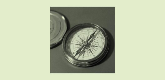 A close-up, black-and-white photograph of an open pocket compass with a decorative wind rose dial, metal casing, and its lid resting beside it on a flat surface. The compass needle points roughly north-northeast.