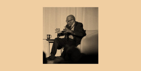 A sepia-toned photograph of an elderly man seated in a white armchair on a stage, gesturing expressively with both hands as he speaks. He is wearing glasses, a dark blazer, and dress pants. A small side table with a dark mug sits beside him. The silhouettes of audience members are visible in the foreground.