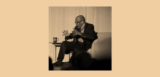 A sepia-toned photograph of an elderly man seated in a white armchair on a stage, gesturing expressively with both hands as he speaks. He is wearing glasses, a dark blazer, and dress pants. A small side table with a dark mug sits beside him. The silhouettes of audience members are visible in the foreground.